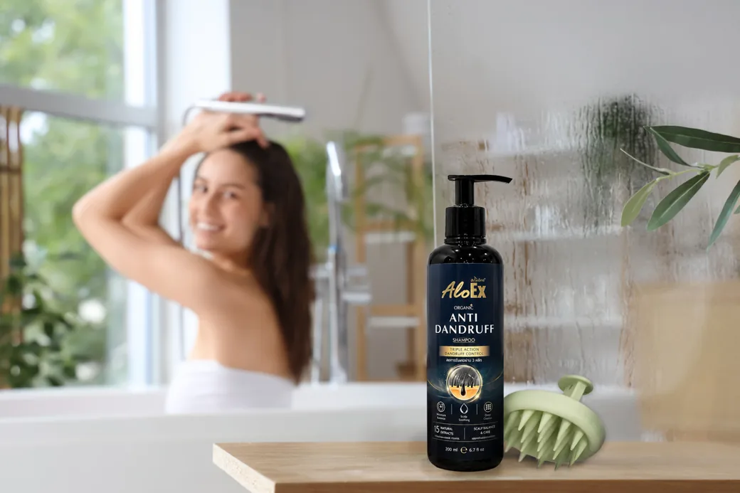 ผู้หญิงกำลังสระผมในห้องน้ำ พร้อมขวดแชมพูขจัดรังแค AloEx Organic Anti-Dandruff Shampoo วางอยู่ข้างอ่าง